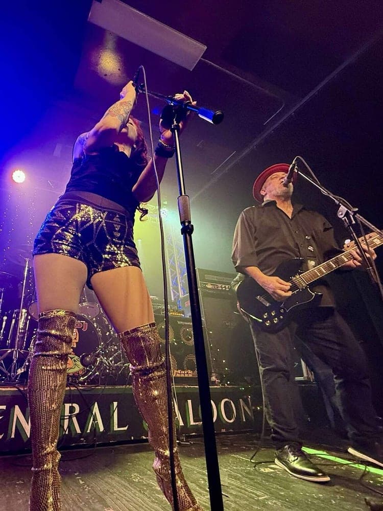 A singer reaching up the mic stand in sequinned boots, beside a guitarist in a red hat.