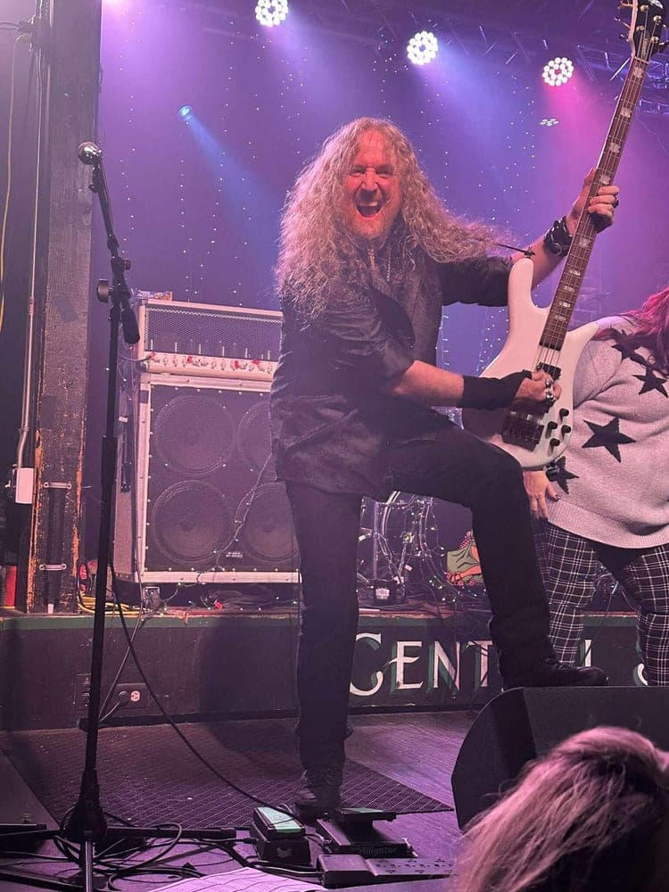 A bassist with long hair grinning wide, one boot up on the monitor, under purple stage lights.